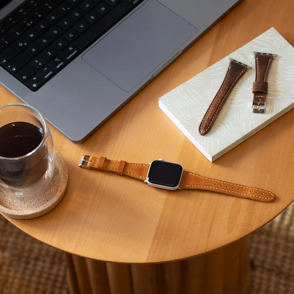 Brown leather Apple Watch strap on a table beside a laptop, coffee, and spare straps.